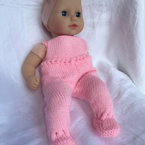 Könnte beinhalten: Ein rosa gestricktes Baby-Puppen-Outfit mit passendem Stirnband. Das Outfit besteht aus einem langärmeligen Oberteil mit Rundhalsausschnitt und einer Hose mit Füssen.