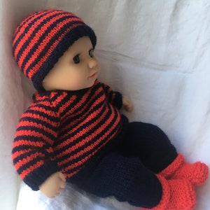May include: A knitted baby doll outfit in red and navy blue stripes. The outfit includes a hat, sweater, trousers, and booties.
