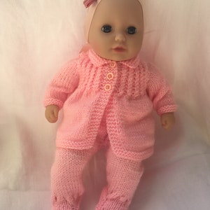 Puede incluir: Una muñeca bebé vestida con un conjunto de punto rosa, que incluye una chaqueta con tres botones, pantalones a juego y una diadema con un lazo. La muñeca tiene ojos azules y una expresión neutra.