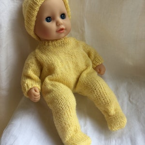 Op de afbeelding: Een gele gebreide babypop outfit met een bijpassende muts. De pop draagt een gele gebreide muts en een gele gebreide romper. De pop heeft blauwe ogen en een lichte huidskleur.
