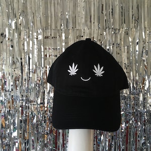 Könnte beinhalten: Schwarze Baseballkappe mit einem weißen lächelnden Gesicht und zwei weißen Marihuana-Blattdesigns über den Augen.