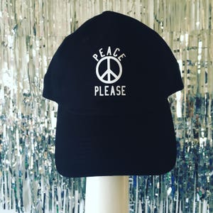 Pode incluir: Um boné de beisebol preto com um símbolo de paz branco e o texto "Peace Please" bordado na frente.