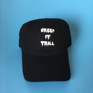 Puede incluir: Gorra de béisbol negra con texto blanco que dice "Creep It Trill" en una fuente que gotea.