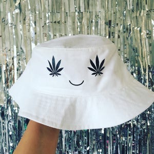 May include: White bucket hat with a black smiley face and two marijuana leaf designs above the smile.