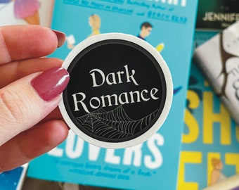 May include: A black and white sticker with the words "Dark Romance" and a spider web design. The sticker is being held by a hand in front of a stack of books with various titles.