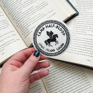 Pu&ograve; includere: Un adesivo rotondo bianco con testo nero e un'illustrazione nera di un cavallo alato. Il testo recita "CAMP HALF-BLOOD" sopra l'immagine e "LONG ISLAND SOUND" sotto. L'adesivo &egrave; tenuto sopra un libro aperto.