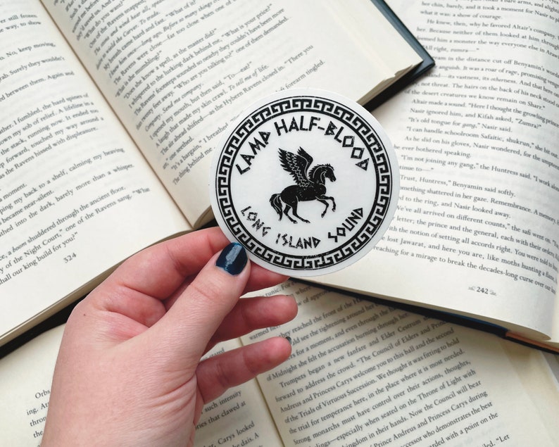 Pu&ograve; includere: Un adesivo rotondo bianco con testo nero e un'illustrazione nera di un cavallo alato. Il testo recita "CAMP HALF-BLOOD" sopra l'immagine e "LONG ISLAND SOUND" sotto. L'adesivo &egrave; tenuto sopra un libro aperto.