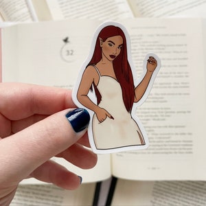 May include: A sticker featuring a stylized illustration of a woman with long auburn hair, wearing a white dress. The woman has pointed ears and is holding her hand up. The sticker is held in front of an open book.