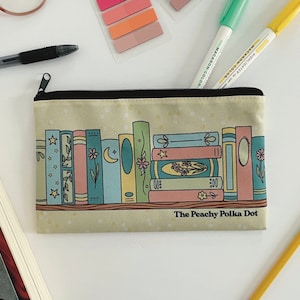 May include: A rectangular pencil case with a black zipper and a beige background. The case features a colorful illustration of a bookshelf with various books. The text "The Peachy Polka Dot" is printed on the case.