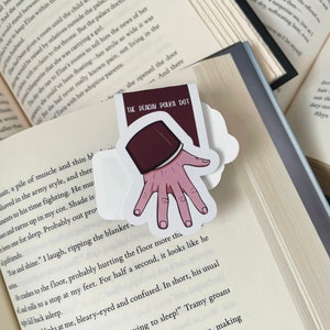 K&ouml;nnte beinhalten: Ein Aufkleber mit einer Hand, die ein Buch mit dem Text "THE PEACUM POLKA DOT" h&auml;lt. Die Hand hat rosa Haut und der Buchumschlag ist weinrot. Der Aufkleber befindet sich auf den Seiten eines offenen Buches.