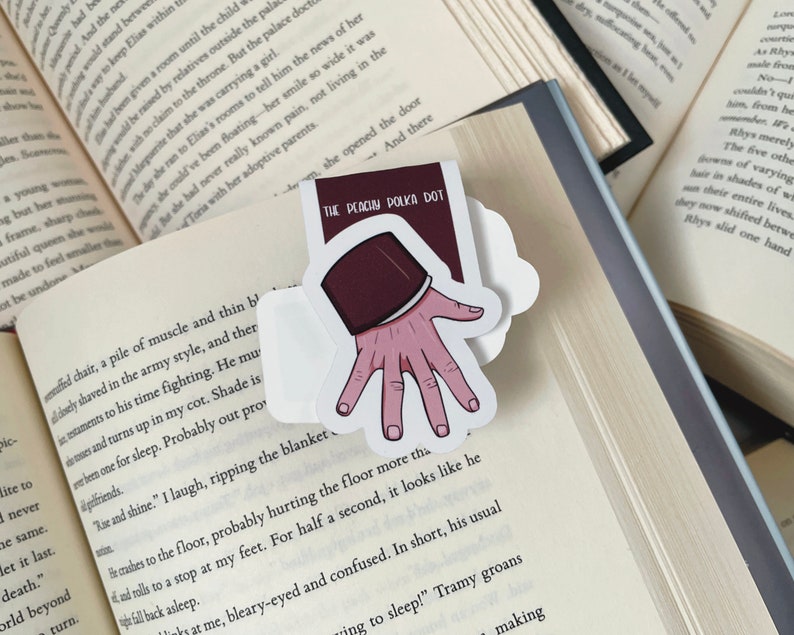 K&ouml;nnte beinhalten: Ein Aufkleber mit einer Hand, die ein Buch mit dem Text "THE PEACUM POLKA DOT" h&auml;lt. Die Hand hat rosa Haut und der Buchumschlag ist weinrot. Der Aufkleber befindet sich auf den Seiten eines offenen Buches.