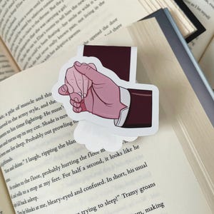 K&ouml;nnte beinhalten: Ein Aufkleber mit einer Handgeste mit rosa Hautton, der eine dunkelburgunderfarbene Anzugjacke tr&auml;gt. Der Aufkleber befindet sich auf einem offenen Buch mit gedrucktem Text. Der Hintergrund umfasst weitere offene B&uuml;cher.