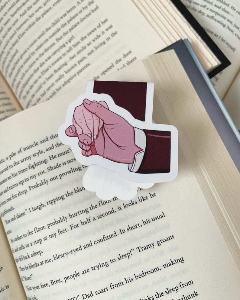 K&ouml;nnte beinhalten: Ein Aufkleber mit einer Handgeste mit rosa Hautton, der eine dunkelburgunderfarbene Anzugjacke tr&auml;gt. Der Aufkleber befindet sich auf einem offenen Buch mit gedrucktem Text. Der Hintergrund umfasst weitere offene B&uuml;cher.