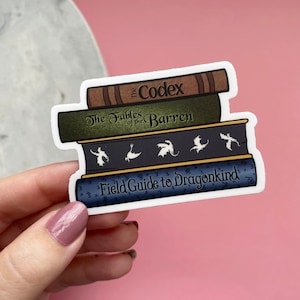 May include: A sticker featuring a stack of fantasy books with titles like "The Codex" and "Field Guide to Dragonkind." The books are in shades of brown, green, and blue, with white outlines. The sticker is held by a hand against a pink background.