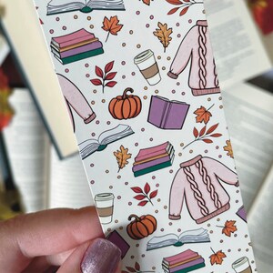 May include: A white bookmark with a colorful pattern of books, coffee cups, pumpkins, and sweaters. The bookmark has a white background with a repeating pattern of these items.