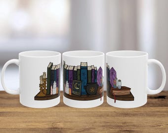 May include: White ceramic mug featuring a bookshelf design with books, crystals, candles, and a cauldron. The design includes a crescent moon and star symbol. The mug is set on a wooden surface.