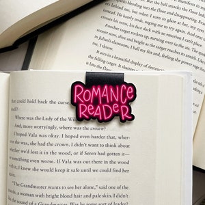 Pu&ograve; includere: Un segnalibro nero e rosa con le parole "Romance Reader" in lettere rosa neon. Il segnalibro &egrave; su un libro con testo su una maledizione e una corona.