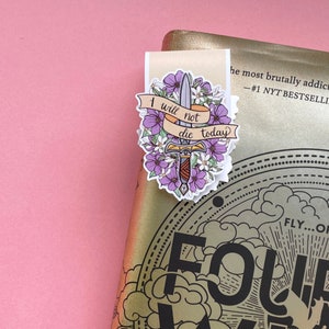May include: A bookmark with a floral and sword design. The bookmark features a banner with the text "I will not die today". The design includes purple flowers and a sword. The bookmark is placed on a gold book.
