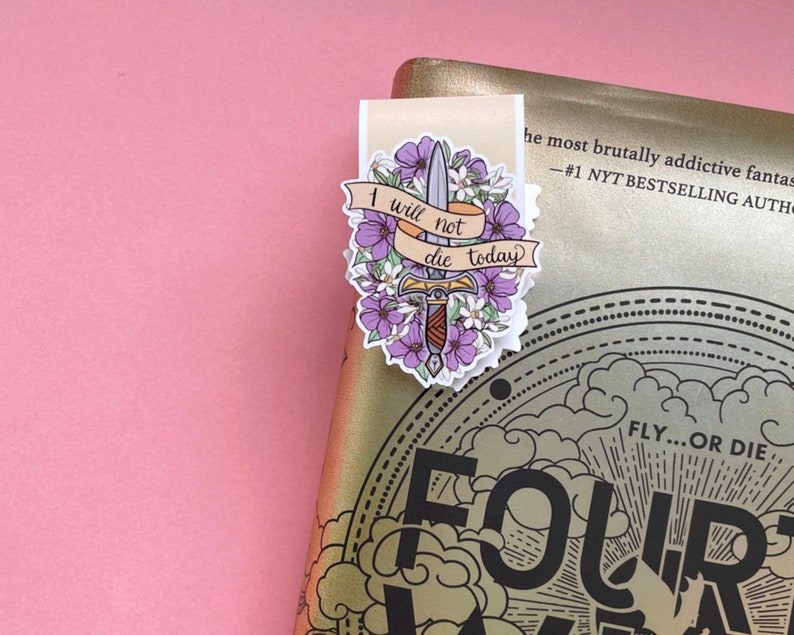 May include: A bookmark with a floral and sword design. The bookmark features a banner with the text "I will not die today". The design includes purple flowers and a sword. The bookmark is placed on a gold book.