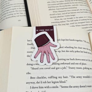 K&ouml;nnte beinhalten: Ein Lesezeichen mit einer Handillustration, mit einem bordeauxfarbenen &Auml;rmel und einer hellrosa Hand mit sichtbaren Fingern&auml;geln. Der Text "THE PEACHY POLKA DOT" ist auf dem Lesezeichen aufgedruckt. Das Lesezeichen befindet sich in einem offenen Buch.