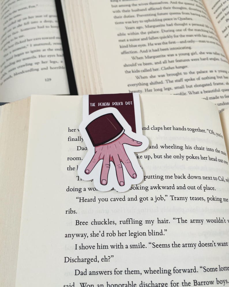 K&ouml;nnte beinhalten: Ein Lesezeichen mit einer Handillustration, mit einem bordeauxfarbenen &Auml;rmel und einer hellrosa Hand mit sichtbaren Fingern&auml;geln. Der Text "THE PEACHY POLKA DOT" ist auf dem Lesezeichen aufgedruckt. Das Lesezeichen befindet sich in einem offenen Buch.
