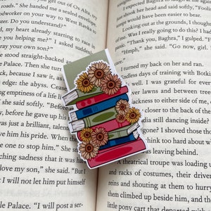 Puede incluir: Un marcapáginas colorido con una pila de libros con un diseño de girasoles. Los libros son en tonos de rojo, azul y verde, con girasoles amarillos en la parte superior. Un accesorio encantador para los amantes de los libros.