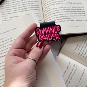Pu&ograve; includere: Un segnalibro nero e rosa con il testo "Romance Reader" in lettere rosa neon. Il segnalibro &egrave; tenuto da una mano con un anello d'oro.