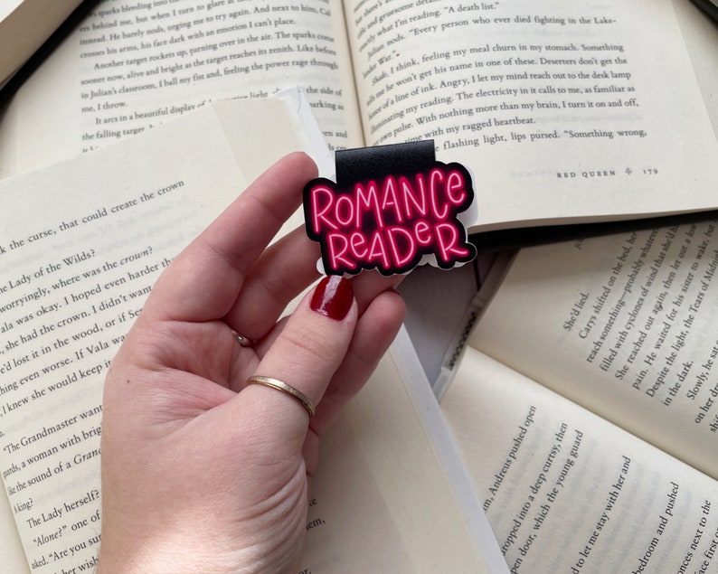 Pu&ograve; includere: Un segnalibro nero e rosa con il testo "Romance Reader" in lettere rosa neon. Il segnalibro &egrave; tenuto da una mano con un anello d'oro.