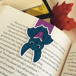 May include: A teal bat bookmark with a purple top, resting on the pages of a book. The bat is upside down with a cute, cartoonish design. A red leaf and other books are in the background.