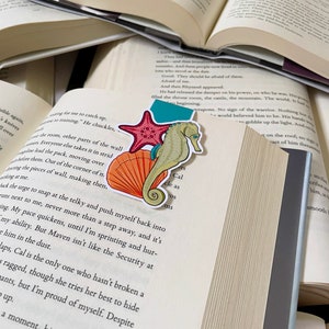 May include: A colorful bookmark featuring a seahorse, a starfish, and a seashell. The bookmark is placed on a page of an open book.