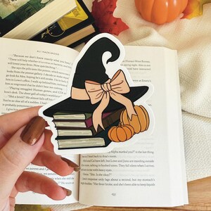 May include: A decorative bookmark with a black witch's hat, peach bow, books, and pumpkins. The bookmark is placed over an open book. Ideal for Halloween or reading enthusiasts.