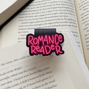 Pu&ograve; includere: Un segnalibro nero e rosa con le parole "Romance Reader" in lettere rosa neon. Il segnalibro ha la forma di un rettangolo con angoli arrotondati.