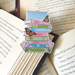 May include: A stack of colourful books with a butterfly on top, surrounded by pink and green flowers. The bookmark is ideal for book lovers and features a whimsical design.