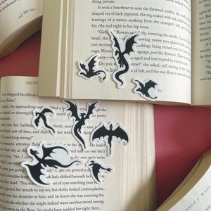 May include: A collection of black dragon silhouette stickers on a book page. The stickers are various sizes and shapes, with some dragons facing forward and others facing sideways. The book page is open and has text printed on it.