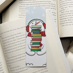 May include: A snowman wearing a red scarf and earmuffs is holding a stack of colorful books. The snowman is on a blue and white background with snowflakes.