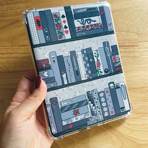 Op de afbeelding: Een doorzichtige beschermhoes voor een e-reader met een boekenplankontwerp. De hoes toont rijen geïllustreerde boeken in grijstinten, met decoratieve elementen zoals rozen, speelkaartsymbolen en een slang. De tekst "Peachy Polka Dot" is zichtbaar.