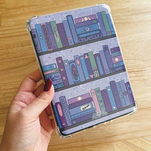 Può includere: Custodia trasparente per telefono con un design stravagante a libreria. La custodia presenta file di libri colorati in tonalità viola, blu e verde, con elementi decorativi come lune, stelle e farfalle. Il testo "Peachy Polka Dot" è visibile.