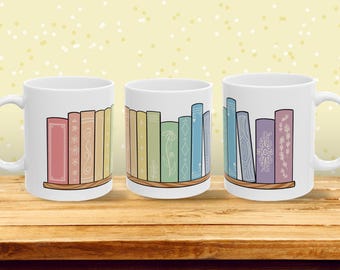 May include: Three white ceramic mugs with a wraparound design of a bookshelf filled with colorful books. The books are in pastel colors and have decorative details. The mugs sit on a wooden surface.