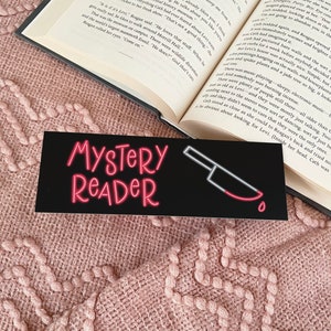 May include: A black bookmark with a red neon sign that says "Mystery Reader" and a red knife with a blood drop. The bookmark is on top of a pink knitted blanket and a book.