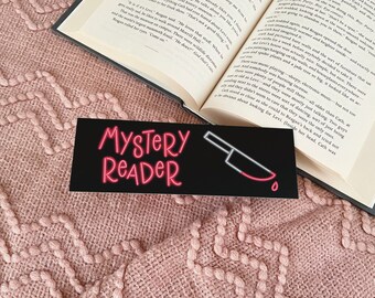 May include: A black bookmark with a red neon sign that says "Mystery Reader" and a red knife with a blood drop. The bookmark is on top of a pink knitted blanket and a book.