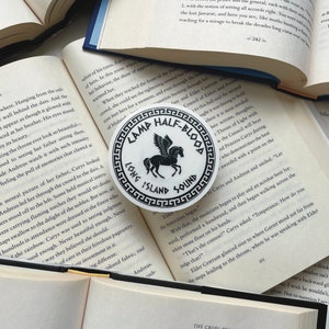 Pu&ograve; includere: Un adesivo rotondo bianco con la scritta nera "CAMP HALF-BLOOD LONG ISLAND SOUND" che circonda un'illustrazione di Pegaso nero. L'adesivo &egrave; posto su un libro aperto.