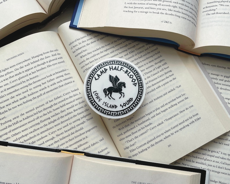 Pu&ograve; includere: Un adesivo rotondo bianco con la scritta nera "CAMP HALF-BLOOD LONG ISLAND SOUND" che circonda un'illustrazione di Pegaso nero. L'adesivo &egrave; posto su un libro aperto.