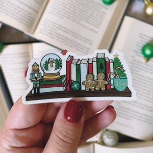 Puede incluir: Una pegatina festiva que representa una estantería con decoraciones navideñas. Incluye una bola de nieve, libros, un árbol de Navidad, hombres de jengibre y una taza con la palabra "joy". La paleta de colores incluye verde, rojo y marrón.