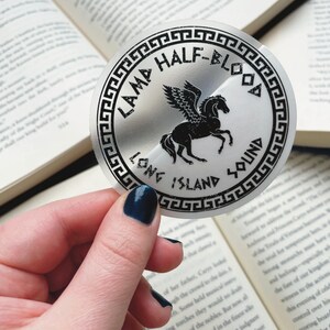 Pu&ograve; includere: Un adesivo circolare con il testo "LAMA HALF-BLOOD LONG ISLAND SOUND" in nero, che circonda un cavallo alato nero. L'adesivo &egrave; tenuto davanti a un libro aperto.