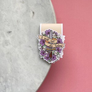 May include: A sticker featuring a sword with a floral design and the text "I will not die today" on a banner. The sticker has purple and white flowers and a beige background.