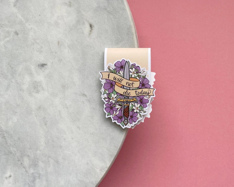 May include: A sticker featuring a sword with a floral design and the text "I will not die today" on a banner. The sticker has purple and white flowers and a beige background.