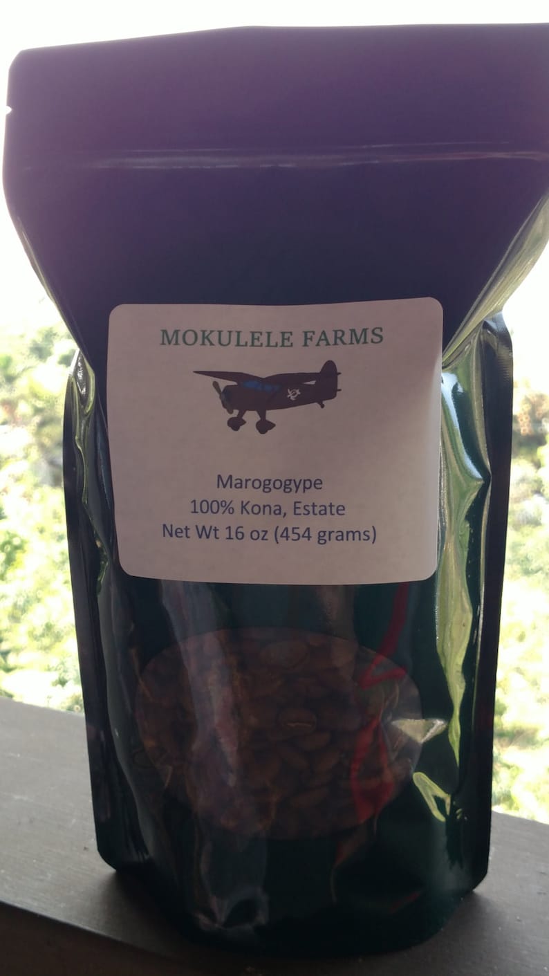 May include: A dark, opaque bag of coffee beans with a label that reads "MOKULELE FARMS." The label features an airplane illustration and text: "Marogogype 100% Kona, Estate Net Wt 16 oz (454 grams)." The bag is partially transparent, revealing the coffee beans.