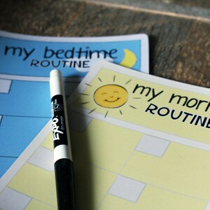 May include: Two dry-erase checklists, one blue with a crescent moon and the text "my bedtime routine", and one yellow with a smiling sun and the text "my morning routine". A black dry-erase marker is lying on top of the checklists.
