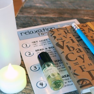 May include: A relaxation tips sheet with four steps listed. The steps include getting cozy, reading a book, using essential oils, and journaling. There is a lit candle, a glass of water, a bottle of essential oil, a notebook, and a blue pencil on the table.