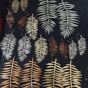 May include: A collection of various metal fern leaf earrings in gold, silver, and copper tones. The earrings feature detailed, elongated leaf shapes with wire hooks for attachment, arranged in multiple sizes and metallic finishes on a dark surface.